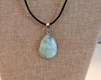 May include: A teardrop-shaped pendant necklace with a light blue stone. The stone is set in a silver-colored metal frame and hangs from a black cord. The necklace is displayed on a tan fabric background.