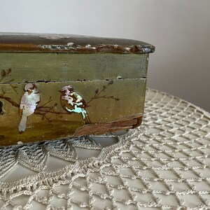 May include: A vintage wooden box with a distressed green finish. The box features two birds with iridescent accents perched on branches. The lid has a dark brown trim. The box is placed on a cream-colored, crocheted doily.