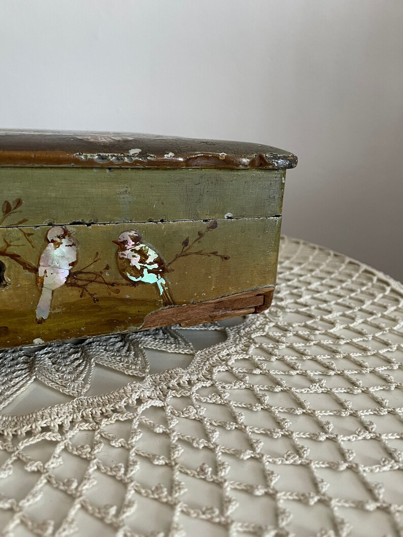May include: A vintage wooden box with a distressed green finish. The box features two birds with iridescent accents perched on branches. The lid has a dark brown trim. The box is placed on a cream-colored, crocheted doily.