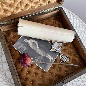 May include: An open antique wooden box with a tufted, gold-colored interior. Inside are a vintage photograph, a dried rose, a rolled paper, and a rhinestone brooch. The box is on a white lace surface.