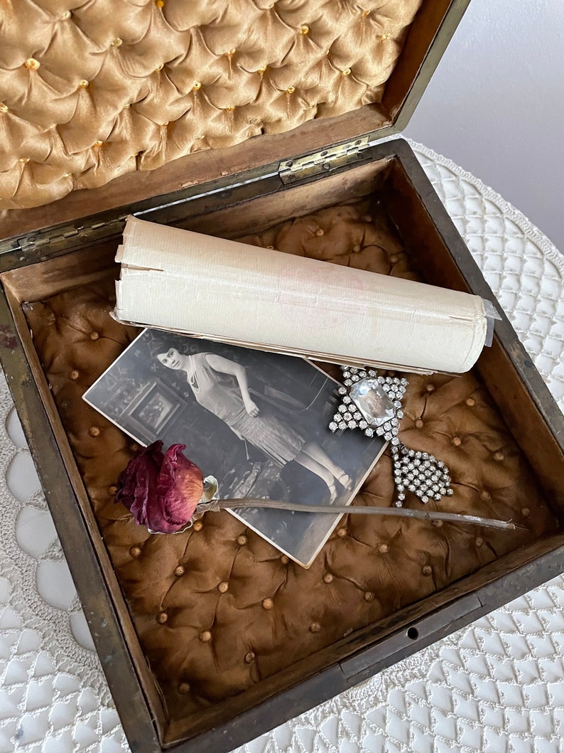 May include: An open antique wooden box with a tufted, gold-colored interior. Inside are a vintage photograph, a dried rose, a rolled paper, and a rhinestone brooch. The box is on a white lace surface.