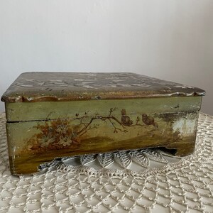 May include: A rectangular, antique wooden box with a hinged lid. The box is painted in shades of green and gold, with a distressed finish. Floral and bird motifs adorn the front. The box sits on a white lace doily.