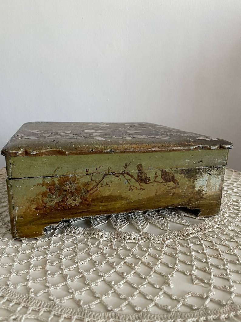 May include: A rectangular, antique wooden box with a hinged lid. The box is painted in shades of green and gold, with a distressed finish. Floral and bird motifs adorn the front. The box sits on a white lace doily.
