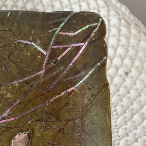 May include: Close-up of a rectangular, antique wooden object with a dark, textured surface. The object features an inlaid design of branches with iridescent accents in pink, green, and blue. The edges are slightly worn.