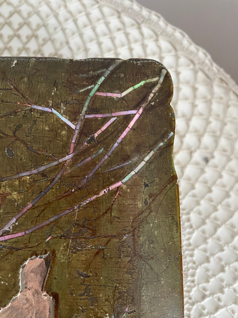 May include: Close-up of a rectangular, antique wooden object with a dark, textured surface. The object features an inlaid design of branches with iridescent accents in pink, green, and blue. The edges are slightly worn.