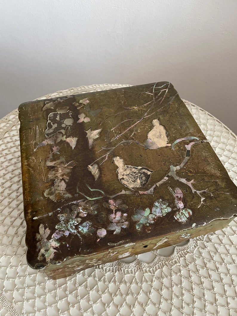 May include: A rectangular, antique wooden box with a detailed, inlaid design. The box features a bird and floral motif with iridescent mother-of-pearl accents. The box has a dark, aged finish and a scalloped edge.