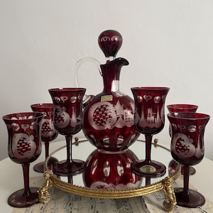 Könnte beinhalten: Ein rubinrotes böhmisches Glas-Dekanter-Set mit sechs passenden Weingläsern. Das Set verfügt über geätzte Designs und steht auf einem verspiegelten Tablett mit goldfarbenem Filigranrand. Die Karaffe hat einen Verschluss.