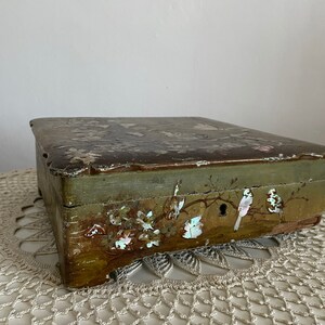 May include: An antique wooden box with a faded green exterior and a decorative lid. The box features painted floral and bird motifs, with mother-of-pearl accents. The box is resting on a cream-colored, crocheted doily.