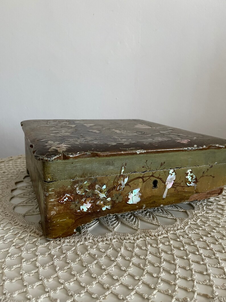 May include: An antique wooden box with a faded green exterior and a decorative lid. The box features painted floral and bird motifs, with mother-of-pearl accents. The box is resting on a cream-colored, crocheted doily.