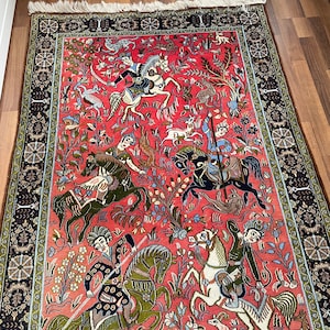 Könnte beinhalten: Ein roter, blauer und grüner persischer Teppich mit einem detaillierten Design von Menschen, die auf Pferden reiten und Tiere jagen. Der Teppich hat einen schwarzen und goldenen Rand.