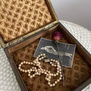May include: An open antique wooden box with a tufted, golden-brown interior. Inside, a pearl necklace, a vintage photograph, and a dried rose are visible. The box has a brass clasp and hinges, suggesting a jewelry or keepsake box.