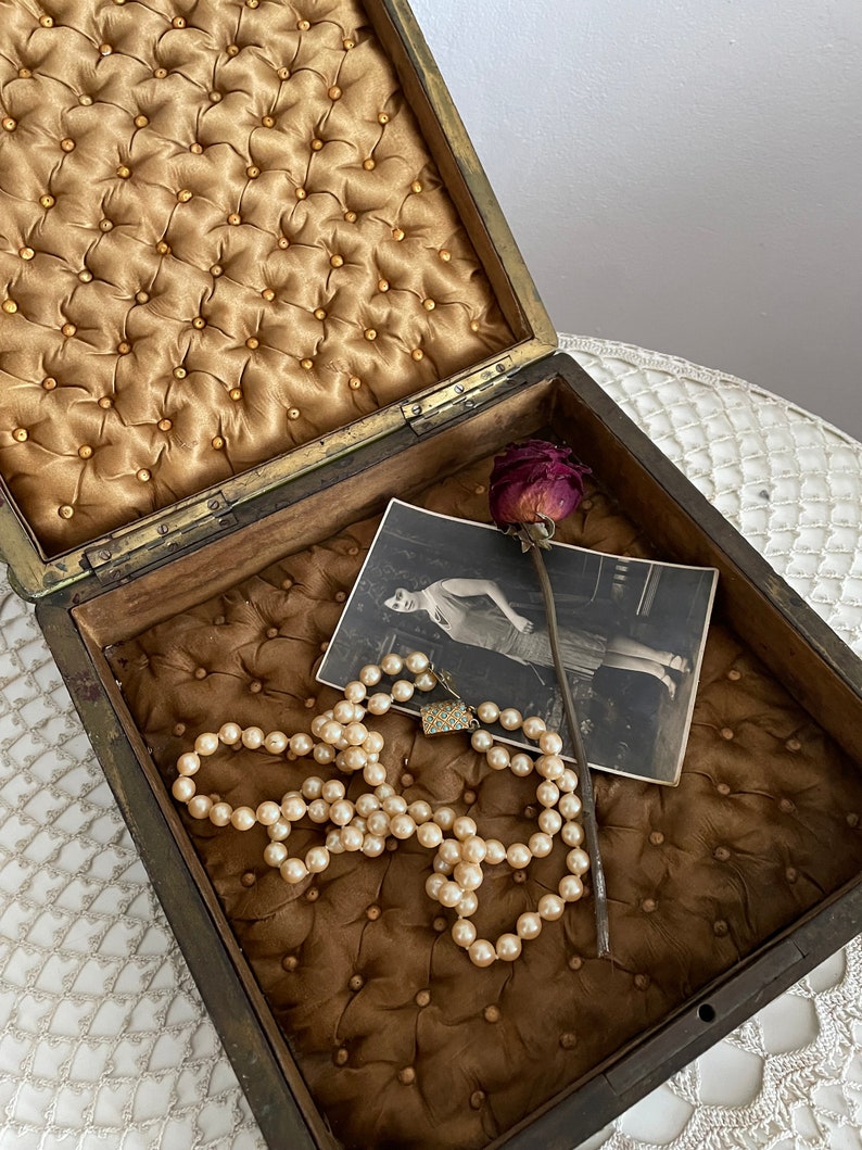 May include: An open antique wooden box with a tufted, golden-brown interior. Inside, a pearl necklace, a vintage photograph, and a dried rose are visible. The box has a brass clasp and hinges, suggesting a jewelry or keepsake box.