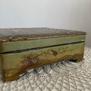 May include: A rectangular, vintage wooden box with a hinged lid. The box is painted in shades of green and brown, with hand-painted floral and bird motifs. The box sits on a white, open-weave surface.