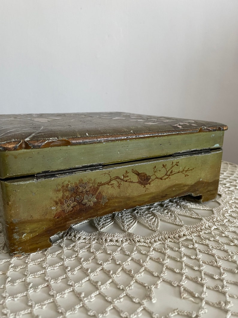 May include: A rectangular, vintage wooden box with a hinged lid. The box is painted in shades of green and brown, with hand-painted floral and bird motifs. The box sits on a white, open-weave surface.