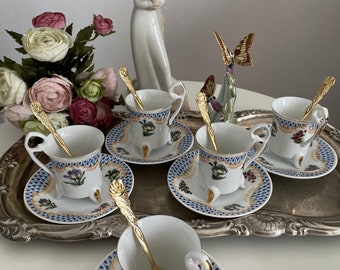 May include: Set of six white porcelain teacups and saucers with gold trim and floral designs. Each cup has three gold feet and comes with a matching gold teaspoon.