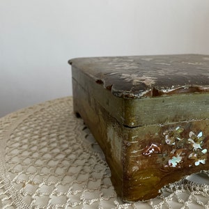 May include: A small, rectangular wooden box with a distressed finish. The box is painted in shades of green and brown, with decorative floral accents. The lid has a scalloped edge.