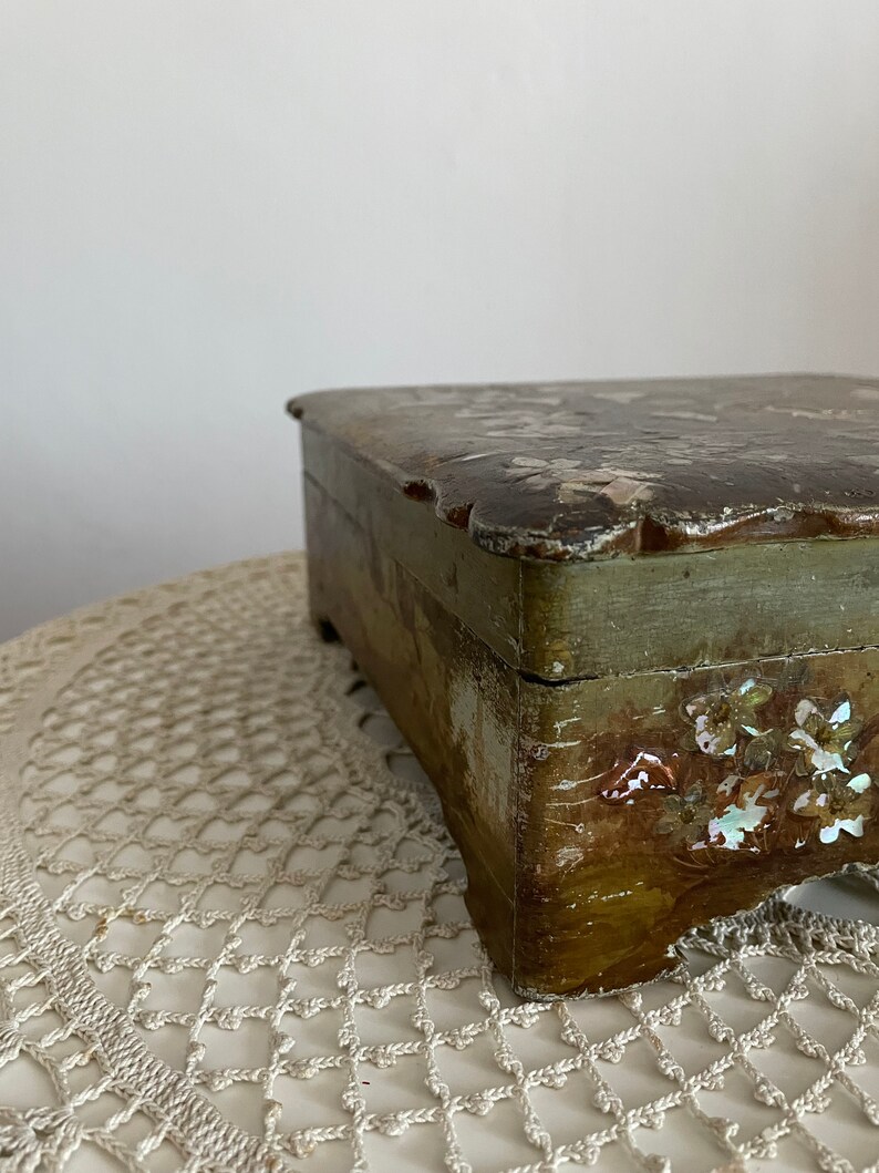 May include: A small, rectangular wooden box with a distressed finish. The box is painted in shades of green and brown, with decorative floral accents. The lid has a scalloped edge.