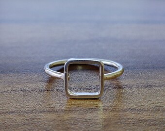 May include: A silver ring with a square-shaped open design. The ring is on a wooden surface.