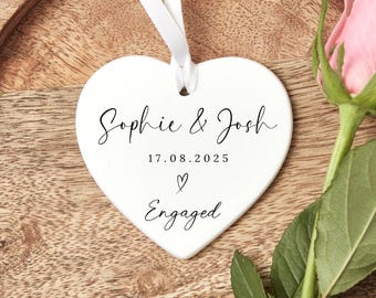 May include: A white heart-shaped ornament with the names "Sophie & Josh", the date "17.08.2025", and the word "Engaged" in black script. A white ribbon is attached for hanging. A pink rose with green leaves is next to the ornament.