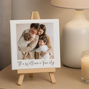 May include: A white ceramic tile with a family photo, displayed on a small wooden easel. The photo features a family of four, with the text "The Williams Family" in script. A lit candle and a lamp are in the background.