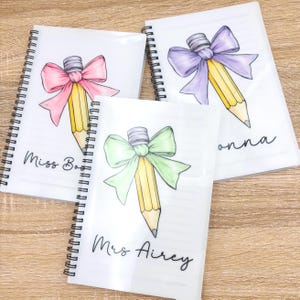 May include: Three spiral bound notebooks with a pencil and bow design. The notebooks are white with black text. The text on the notebooks reads "Miss Bao", "Mrs Airey", and "Donna".