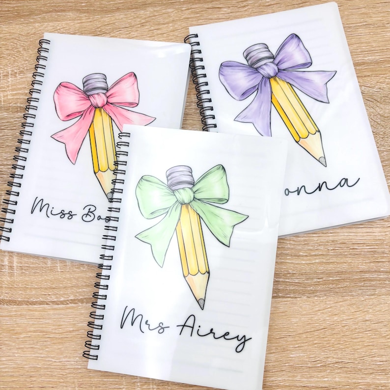 May include: Three spiral bound notebooks with a pencil and bow design. The notebooks are white with black text. The text on the notebooks reads "Miss Bao", "Mrs Airey", and "Donna".