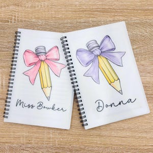 May include: Two white spiral notebooks with personalized names, "Miss Bowker" and "Donna". Each notebook features a yellow pencil with a pink and purple bow, respectively.