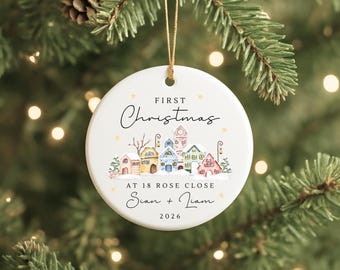 May include: A round white ceramic ornament hanging by a gold string. The design features a colorful illustration of a snowy village with houses, the text "FIRST Christmas AT 18 ROSE CLOSE Sian + Liam 2026," and small gold stars scattered around.