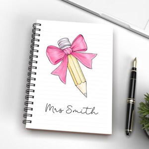 May include: A spiral bound notebook with a pink bow tied around a yellow pencil. The notebook has the name "Mrs. Smith" written on the front.