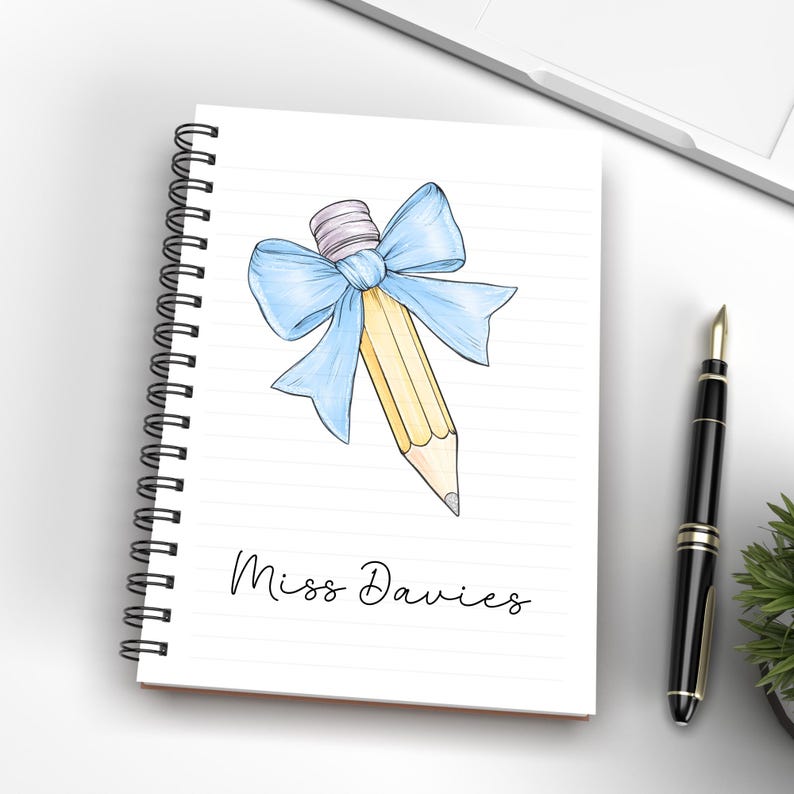 May include: A spiral bound notebook with a yellow pencil tied with a blue bow. The notebook has the name "Miss Davies" written on the front.