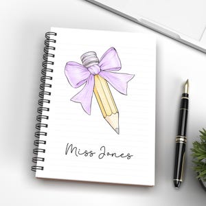 May include: A spiral bound notebook with a pencil tied with a purple bow. The notebook has lined paper and the text "Miss Jones" is written on the front.
