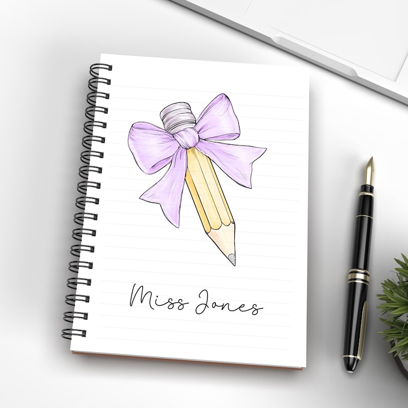 May include: A spiral bound notebook with a pencil tied with a purple bow. The notebook has lined paper and the text "Miss Jones" is written on the front.