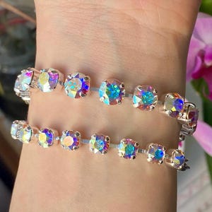 Op de afbeelding: Twee iriserende strass armbanden. Elke armband heeft een reeks ronde, kleurrijke strass-steentjes gezet in een zilverkleurig metaal. De strass-steentjes reflecteren een regenboog aan kleuren, waardoor een sprankelend effect ontstaat. De armbanden worden om een pols gedragen.