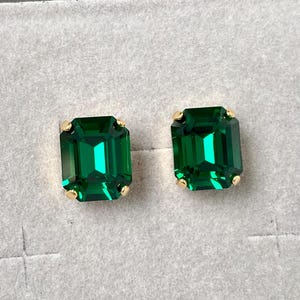 May include: A pair of emerald-cut, vibrant green stud earrings. Each earring features a large, rectangular green stone set in a gold-coloured metal. The earrings are displayed on a light grey surface.