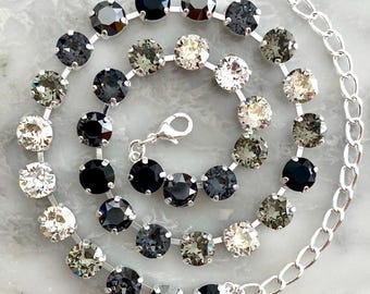 Puede incluir: Un collar plateado con una serie de piedras preciosas redondas y facetadas en tonos negros, grises y claros. El collar está dispuesto en un patrón en espiral, con una cadena y un cierre.
