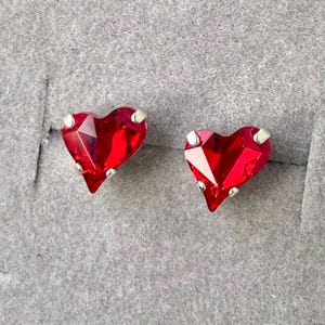 Peut inclure: Une paire de boucles d'oreilles à clous en forme de cœur. Chaque boucle d'oreille présente une pierre précieuse rouge vif à facettes sertie dans un métal de couleur argentée. Les boucles d'oreilles sont présentées sur un fond gris.