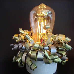 May include: A decorative lamp with a large, clear glass light bulb and a base made of concrete. The base is surrounded by a cluster of gold and silver metallic ribbon.