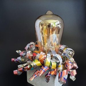 May include: A light bulb with a gold finish is surrounded by colorful, twisted aluminum foil. The bulb is sitting on a square, concrete base.