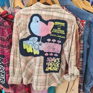 May include: A beige and pink plaid shirt with a large patch on the back. The patch features a bird, guitar, and text that reads "Woodstock Music & Art Fair" and "3 Days of Peace & Music". The shirt is displayed with other clothing.