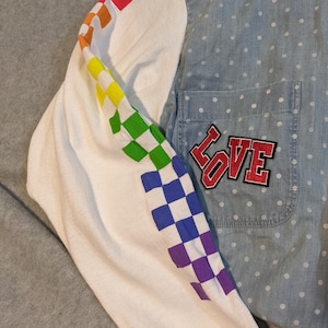 May include: A white long-sleeved shirt with a rainbow checkered pattern on the sleeve. The shirt has a denim blue button-up collar and a pocket with a red "LOVE" patch on the right side.