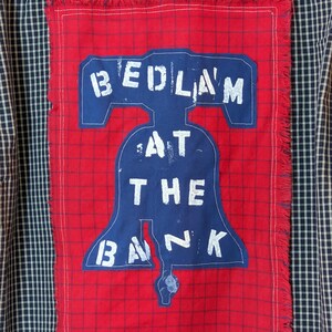 May include: A red and white plaid fabric patch with a blue Liberty Bell design. The patch has the text "BEDLAM AT THE BANK" written on it.