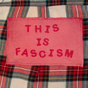 May include: A red rectangular patch with the words "THIS IS FASCISM" in bold, pink letters. The patch is sewn onto a plaid fabric with red, black, green, and white stripes.