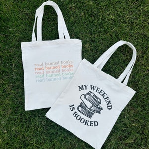 May include: Two white canvas tote bags. One bag has the text "read banned books" repeated five times in different colors. The other bag has the text "MY WEEKEND IS BOOKED" with a stack of books illustrated below.