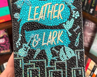 May include: A book cover for "Leather & Lark" from "The Ruinous Love Trilogy." The cover is black, embellished with small, sparkling black and turquoise gems. The title is in gold lettering, with turquoise accents. The book is displayed on a bookshelf.
