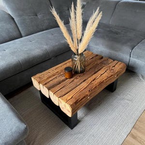Puede incluir: Una mesa de centro rectangular construida con vigas de madera gruesas en tonos naturales con vetas y texturas visibles. La mesa cuenta con una base de metal negro resistente y está decorada con un jarrón de cristal con hierba de la pampa seca.