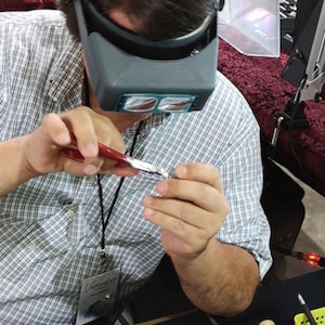 Könnte beinhalten: Eine Person in einem blau-weiß karierten Hemd verwendet eine Zange, um an einem kleinen Objekt zu arbeiten. Sie trägt eine Lupenbrille und ein Lanyard mit einem Ausweis, auf dem "Gemstones" steht.