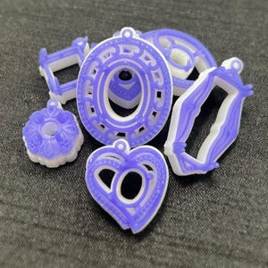 Op de afbeelding: Een set van zes paarse en witte plastic hangerbedels in verschillende vormen, waaronder een hart, een cirkel, een vierkant en een bloem.