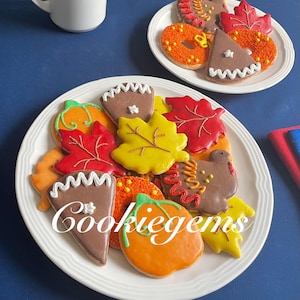 May include: Plates of decorated fall-themed cookies. The cookies are shaped like pumpkins, leaves, and turkey. The cookies are decorated with orange, red, yellow, and brown icing. The text "Cookiegems" is on the plate.