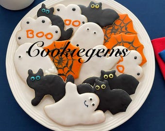 May include: A white plate filled with assorted Halloween-themed sugar cookies, including white ghosts, black cats, black bats, and orange spider webs. Some cookies feature the text "BOO" in orange icing. The cookies are decorated with colorful icing.