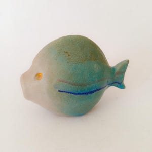 Op de afbeelding: Een keramische vissculptuur met een afgerond lichaam en een kleine staartvin. De vis heeft een lichtgrijze kop en een teal lichaam met blauwe en grijze strepen. Het oog is een kleine, gele stip.